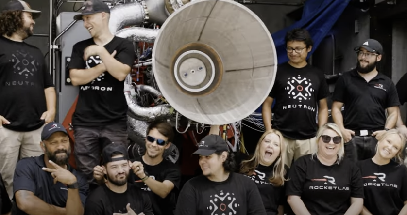 Rocket Lab team members smiling and posing in front of a rocket engine, demonstrating a collaborative and enthusiastic work environmen